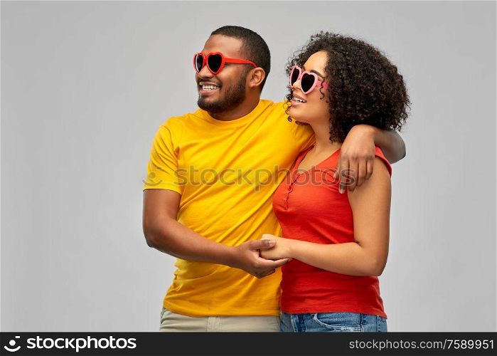 love, relationships and valentines day concept - happy smiling african american couple in heart shaped sunglasses looking at something over grey background. happy african couple in heart shaped sunglasses