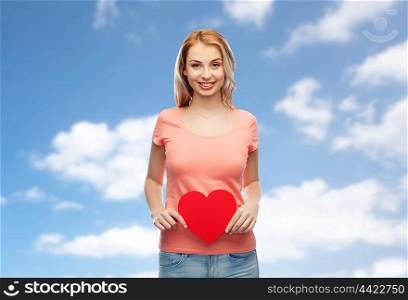 love, romance, charity, valentines day and people concept - smiling young woman or teenage girl with blank red heart shape over blue sky and clouds background