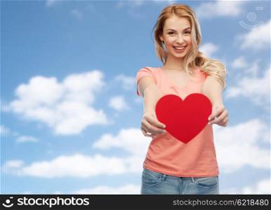 love, romance, charity, valentines day and people concept - smiling young woman or teenage girl with blank red heart shape over blue sky and clouds background