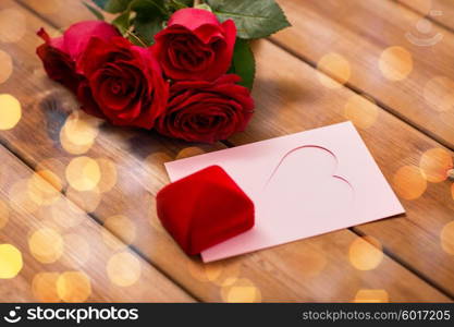 love, romance, valentines day and holidays concept - close up of gift box, red roses and greeting card with heart and golden lights on wood