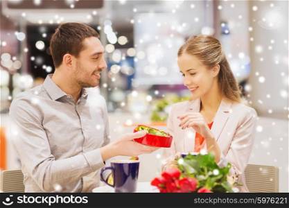 love, romance, valentines day, couple and people concept - happy young couple with red flowers and open chocolate box in at cafe mall with snow effect