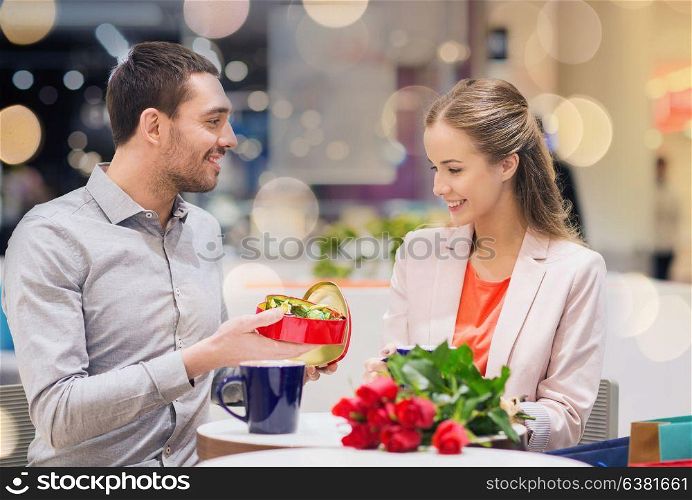 love, romance, valentines day, couple and people concept - happy young couple with red flowers and open gift box in at cafe mall. happy couple with present and flowers in mall