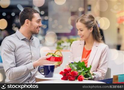 love, romance, valentines day, couple and people concept - happy young couple with red flowers and open gift box in at cafe mall. happy couple with present and flowers in mall