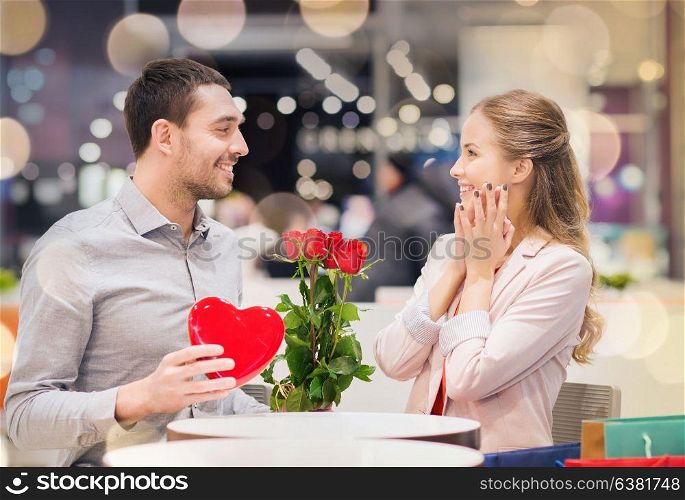 love, romance, valentines day, couple and people concept - happy young man with red flowers giving present to smiling woman at cafe in mall. happy couple with present and flowers in mall
