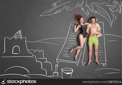 Love story concept of a romantic couple against chalk drawings background. Male lying on sun lounger, wearing headphones and reading a book, female taking picture of a sand castle.