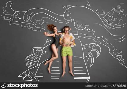 Love story concept of a romantic couple against chalk drawings background. Male lying on sun lounger, wearing headphones and reading a book, female taking selfie with a smartphone.