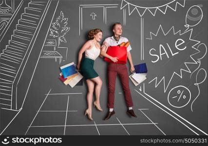 Love story concept of a romantic couple on shopping against chalk drawings background. Young happy couple standing together with shopping bags in a shopping mall during sales.