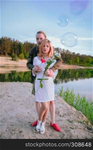 Love story - young happy couple keeps flowers and red heart among soap bubbles by the lake at sunny summer day. Selective focus.. Loving Couple On The Lakeshore
