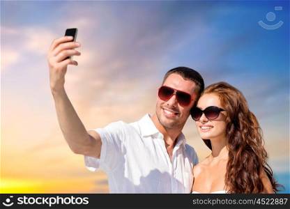 love, summer and technology concept - smiling couple in sunglasses making selfie by smartphone over evening sky background. smiling couple making selfie by smartphone. smiling couple making selfie by smartphone