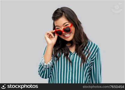 love, summer and valentines day concept - happy asian young woman in heart shaped sunglasses over grey background. happy asian woman with in heart shaped sunglasses