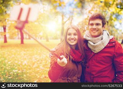 love, technology, relationship, family and people concept - happy smiling couple taking picture by smartphone selfie stick in autumn park. couple taking selfie by smartphone in autumn park