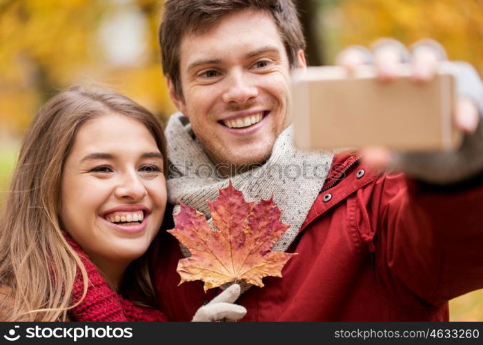 love, technology, relationship, family and people concept - smiling couple with maple leaf taking selfie by smartphone in autumn park