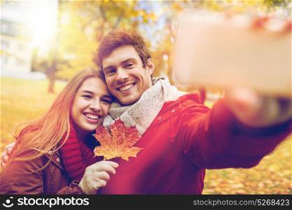 love, technology, relationship, family and people concept - smiling couple with maple leaf taking selfie by smartphone in autumn park. couple taking selfie by smartphone in autumn park