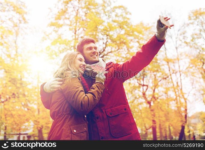 love, technology, relationship, family and people concept - smiling couple with maple leaf taking selfie by smartphone in autumn park. couple taking selfie by smartphone in autumn park