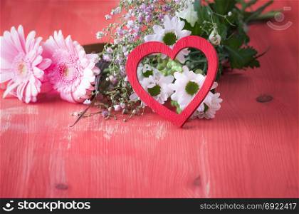 Love theme image with a red wooden heart propped against a bouquet of flowers, on a red wooden table. A concept for relationship, love, valentine day, greeting card.