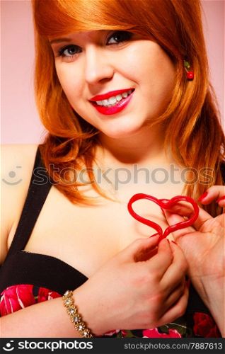Love to candy and sweets. Valentine&#39;s Day concept. Gorgeous young red haired woman holding candy heart on pink background in studio.