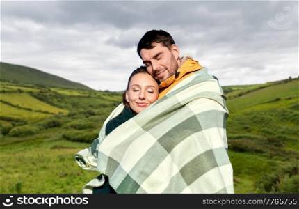 love, travel and tourism concept - happy smiling couple in warm blanket over farmland fields and hills at wild atlantic way in ireland background. happy couple in warm blanket in ireland