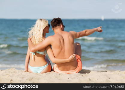 love, travel, tourism and people concept - smiling couple on vacation in swimwear sitting, hugging and pointing finger on summer beach from back