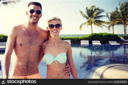 love, travel, tourism, summer and people concept - smiling couple on vacation in swimwear and sunglasses hugging over hotel resort and swimming pool background