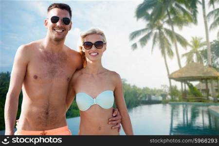love, travel, tourism, summer and people concept - smiling couple on vacation in swimwear and sunglasses hugging over hotel resort and swimming pool background