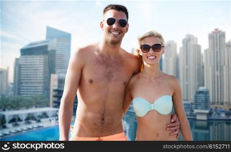 love, travel, tourism, summer and people concept - smiling couple on vacation in swimwear and sunglasses hugging over dubai city waterfront background