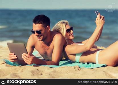 love, travel, tourism, technology and people concept - happy couple on vacation with tablet pc computer and smartphone sunbathing on summer beach