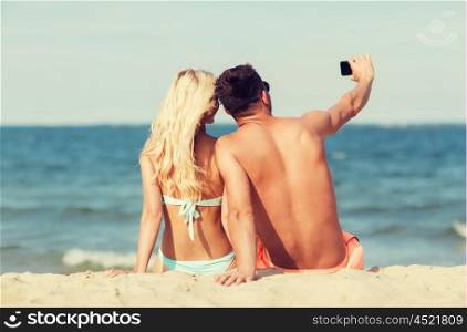 love, travel, tourism, technology and people concept - smiling couple on vacation in swimwear sitting on summer beach and taking selfie with smartphone from back