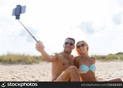 love, travel, tourism, technology and people concept - smiling couple on vacation in swimwear sitting on summer beach and taking picture with smartphone selfie stick