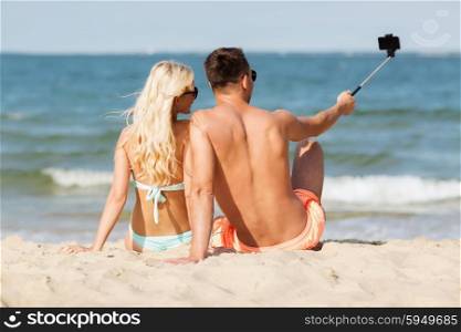 love, travel, tourism, technology and people concept - smiling couple on vacation in swimwear sitting on summer beach and taking picture with smartphone selfie stick from back
