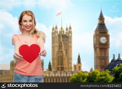 love, travel, tourism, valentines day and people concept - smiling young woman or teenage girl with blank red heart shape over big ben london and city background