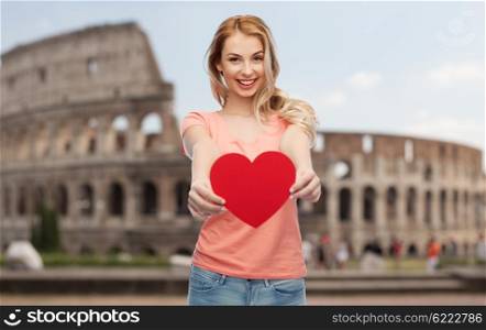 love, travel, tourism, valentines day and people concept - smiling young woman or teenage girl with blank red heart shape over coliseum background