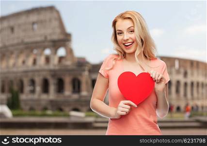 love, travel, tourism, valentines day and people concept - smiling young woman or teenage girl with blank red heart shape over coliseum background