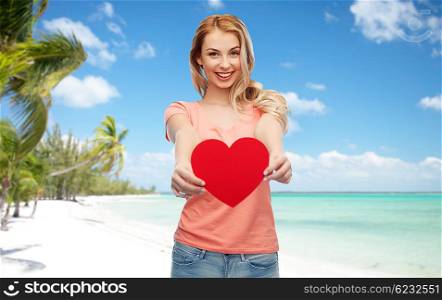 love, travel, tourism, valentines day and people concept - smiling young woman or teenage girl with blank red heart shape over exotic tropical beach with palm trees background