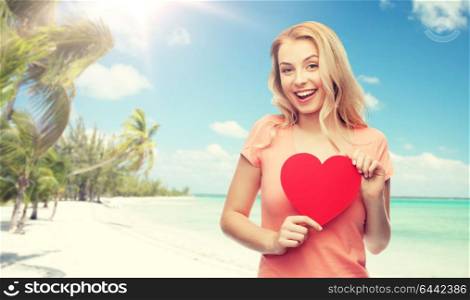 love, travel, tourism, valentines day and people concept - smiling young woman or teenage girl with blank red heart shape over exotic tropical beach with palm trees background. happy woman or teen girl with red heart shape