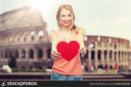 love, travel, tourism, valentines day and people concept - smiling young woman or teenage girl with blank red heart shape over coliseum background. happy woman or teen girl with red heart shape