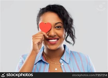 love, valentine&rsquo;s day and charity concept - happy african american young woman covering one eye with red heart over grey background. happy african american woman with red heart