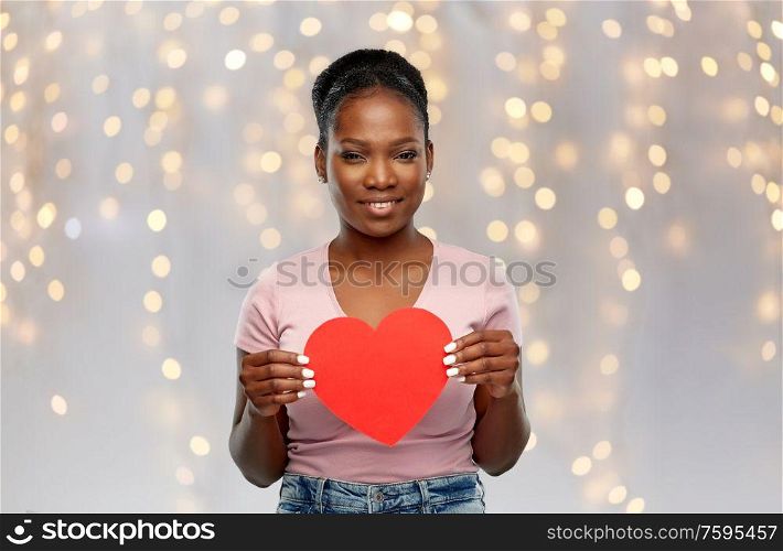 love, valentine&rsquo;s day and charity concept - happy african american young woman with red heart over festive lights background. happy african american woman with red heart