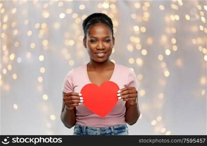 love, valentine&rsquo;s day and charity concept - happy african american young woman with red heart over festive lights background. happy african american woman with red heart