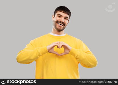 love, valentine&rsquo;s day and people concept - smiling young man in yellow sweatshirt girl making hand heart gesture over grey background. smiling young man in yellow sweatshirt