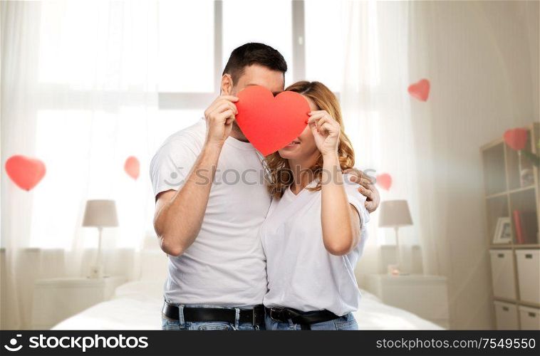 love, valentine&rsquo;s day and relationships concept - smiling couple hiding behind big red heart over bedroom decorated with balloons background. couple hiding behind red heart on valentines day
