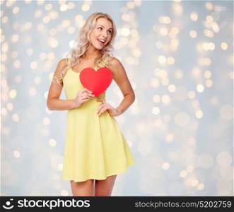 love, valentines day and people concept - smiling young woman in dress with red paper heart over holidays lights background. happy young woman with red paper heart