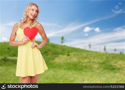 love, valentines day and people concept - smiling young woman in summer dress with red paper heart over blue sky and green field background. happy young woman in summer dress with red heart