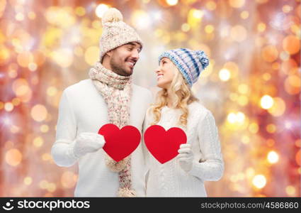love, valentines day, couple, christmas and people concept - smiling man and woman in winter hats and scarf holding red paper heart shape over holidays lights background