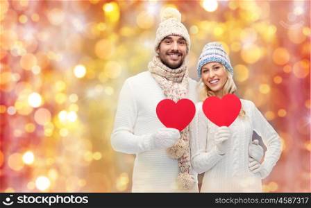 love, valentines day, couple, christmas and people concept - smiling man and woman in winter hats and scarf holding red paper heart shapes over lights background