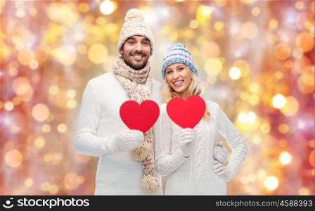 love, valentines day, couple, christmas and people concept - smiling man and woman in winter hats and scarf holding red paper heart shape over holidays lights background