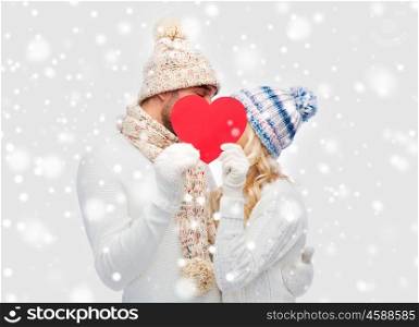 love, valentines day, couple, christmas and people concept - smiling man and woman in winter hats and scarf hiding behind red paper heart shape
