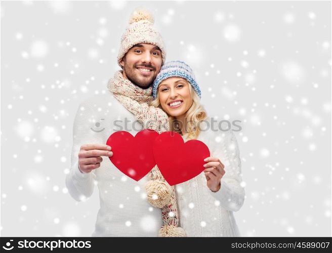 love, valentines day, couple, christmas and people concept - smiling man and woman in winter hats and scarf holding red paper heart shapes
