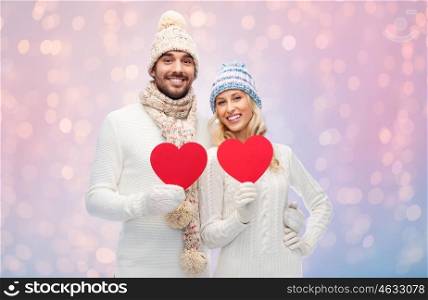 love, valentines day, couple, christmas and people concept - smiling man and woman in winter hats and scarf holding red paper heart shapes over rose quartz and serenity lights background