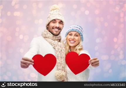 love, valentines day, couple, christmas and people concept - smiling man and woman in winter hats and scarf holding red paper heart shapes over rose quartz and serenity lights background