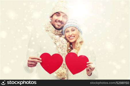 love, valentines day, couple, christmas and people concept - smiling man and woman in winter hats and scarf holding red paper heart shapes. smiling couple in winter clothes with red hearts